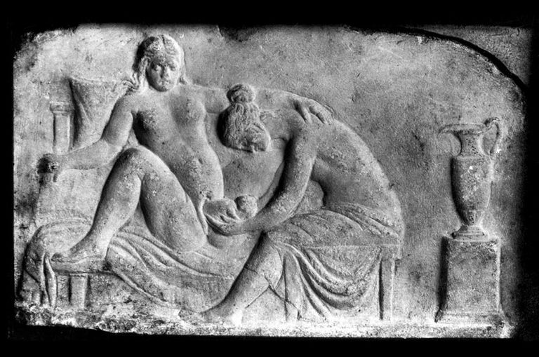 Ancient Greek relief sculpture depicting two female figures in a reclining position, with one figure cradling a small object and the other seated beside her.